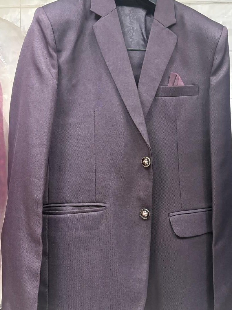 A single-breasted blazer in a muted plum purple color, featuring notch lapels, two front buttons, flap pockets, and a matching pocket square, displayed on a hanger against a tiled background.