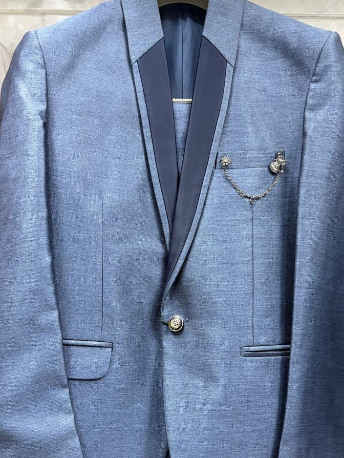 A light blue textured suit jacket with dark navy satin peak lapels, displayed on a hanger. The jacket features a single button closure, flap pockets, and decorative silver-toned lapel pins.