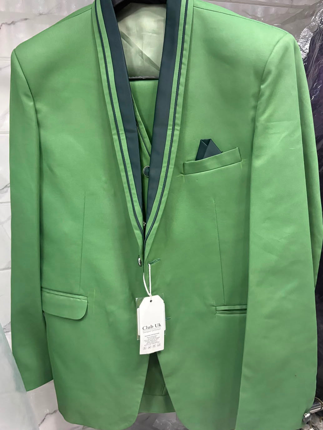 A green men's suit jacket with dark green satin peak lapels, hanging on a black hanger against a light gray marble-patterned wall. A matching green vest and trousers are partially visible underneath, along with a white price tag and a dark green pocket square.