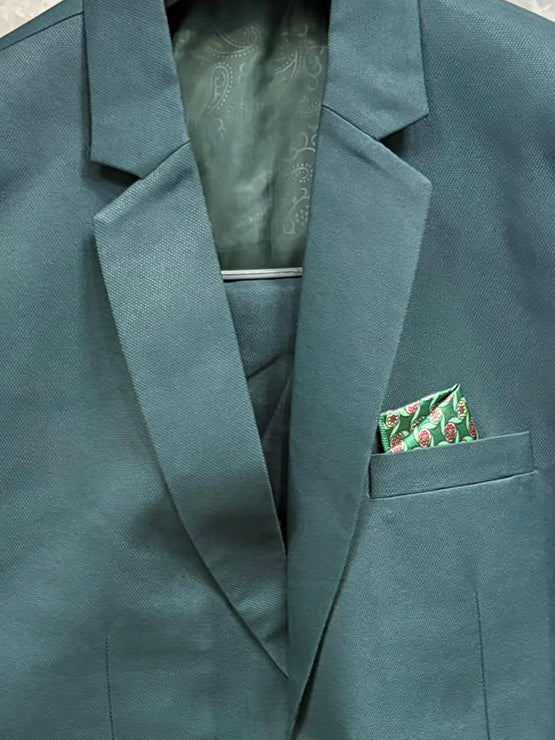 A dark teal-green men's blazer jacket with notch lapels, two buttons, and a patterned pocket square, shown hanging on a hanger against a tiled wall with plastic wrapping.