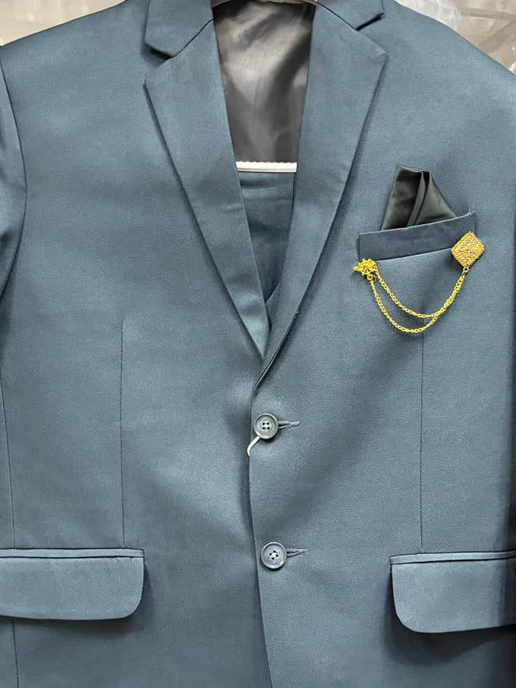 A blue-gray formal suit jacket with matching trousers, a pocket square, and a gold chain detail, hanging on a hanger with plastic wrapping. A purple garment is partially visible on the right.