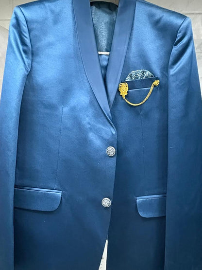 A blue satin blazer jacket with notched lapels, two silver buttons, flap pockets, and a decorative gold and blue pocket square, hanging on a rack.