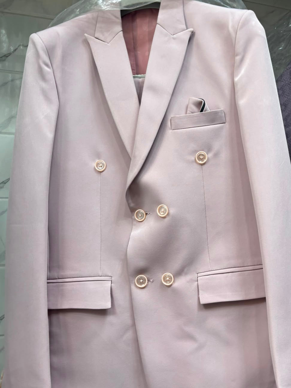 A light pink double-breasted blazer jacket with gold buttons, displayed on a hanger under a clear plastic cover.