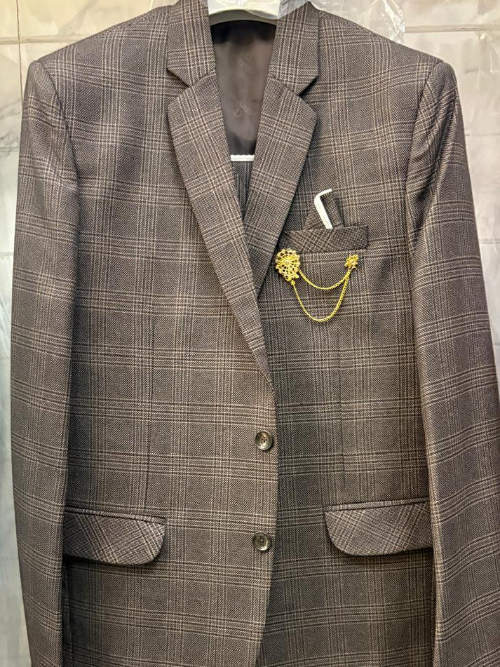 A brown and gray plaid blazer jacket hanging on a hanger, featuring a gold chain brooch pinned to the chest pocket. The jacket is displayed in a retail or storage setting with other garments visible in the background.