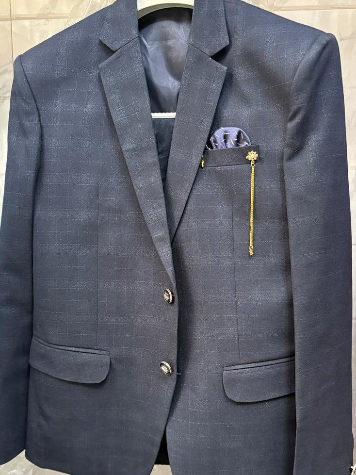 A dark blue-gray plaid blazer jacket with a matching pocket square and a gold lapel pin, hanging on a white hanger on a clothing rack.